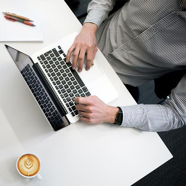 Person typing on a laptop, at a white desk. There is a coffee on the desk also.