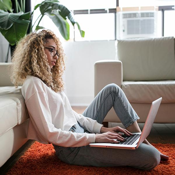 Woman sat on the floor, leaning against a sofa looking at her laptop